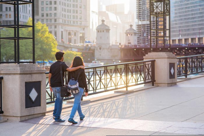 date night chicago image of Beautiful couple goes for a walk at sunset along Riverwalk in Chicago, with skyscrapers and the Chicago River beyond.