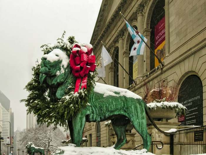 A bronze lion statue wearing a festive wreath with a red bow stands in front of the snow-covered Art Institute of Chicago building